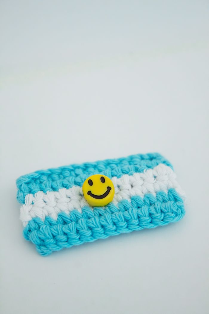 A crocheted wallet in blue and white hues with a cheerful smiley face button.